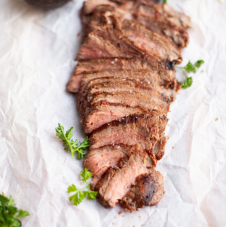 Sliced rosemary garlic flank steak garnished with fresh parsley on parchment paper, with half an avocado in the background.