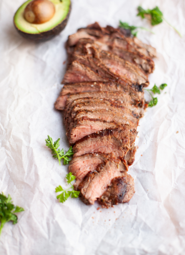 Sliced rosemary garlic flank steak garnished with fresh parsley on parchment paper, with half an avocado in the background.