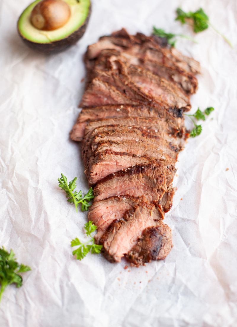 Sliced rosemary garlic flank steak garnished with fresh parsley on parchment paper, with half an avocado in the background.