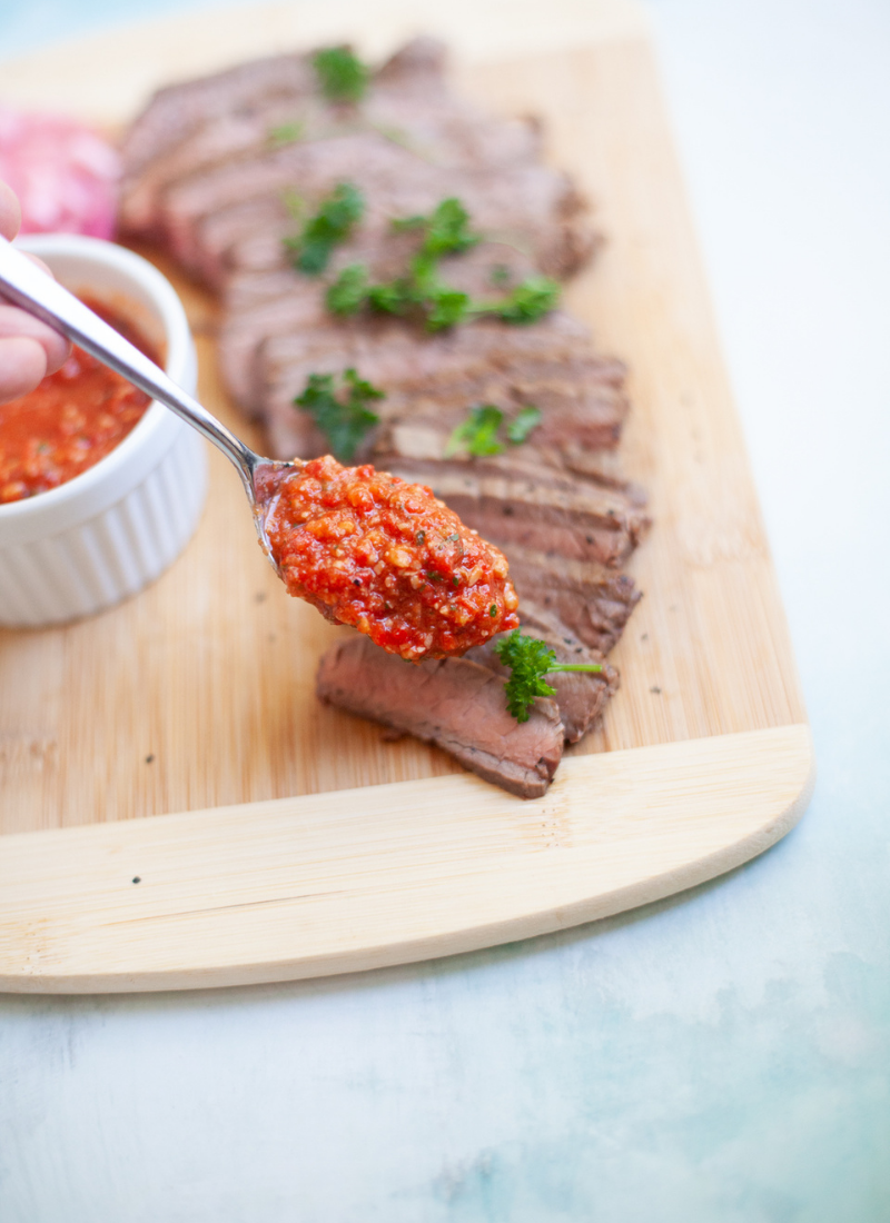 Romesco sauce on a metal spoon over flank steak on a wooden cutting board.