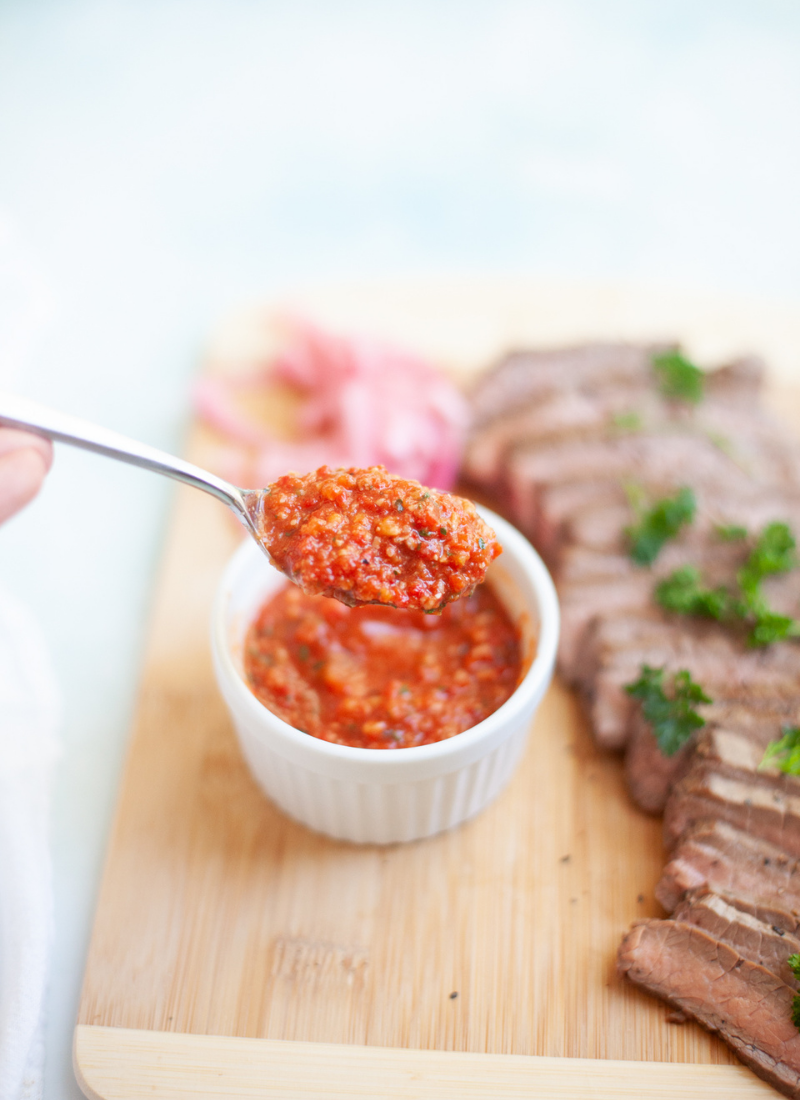 Romesco sauce on a metal spoon above a wooden cutting board with flank steak.