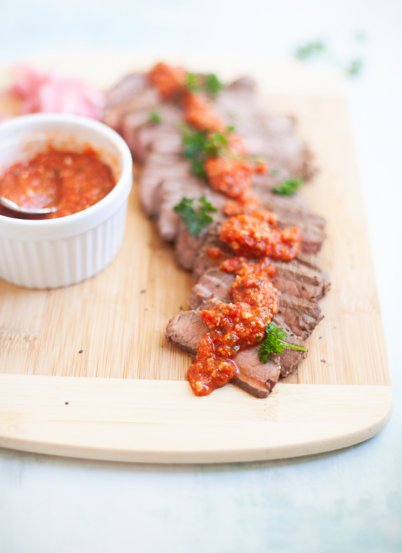 Romesco Sauce drizzled over the top of flank steak on a cutting board. A small bowl of Romesco sitting off to the side.