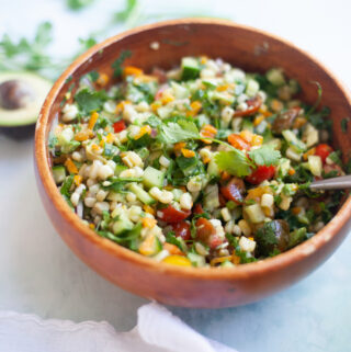 A wooden bowl filled with colorful chopped salad, including corn, tomatoes, cilantro, and cucumber—reminiscent of a fresh and easy corn salsa—sits on a light surface with a spoon and a white cloth nearby.
