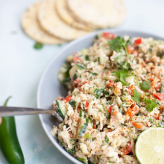 A bowl of creamy Quick Mexican Chicken Salad with chopped herbs and vegetables, garnished with a lime wedge. Sliced crackers and a green chili pepper are beside the bowl. A spoon is scooping some salad.