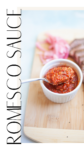 A ramekin of vibrant Romesco sauce sits on a wooden board, with a spoon lifting some out. Pickled onions and part of a grilled meat dish are visible in the background. The text ROMESCO SAUCE is along the left side.