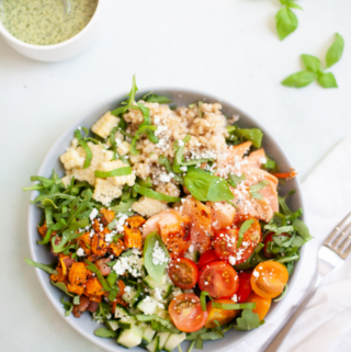 A bowl of salmon sweet potato and chimichurri salad featuring fresh greens, cherry tomatoes, sweet potatoes, corn, grains, salmon, and feta cheese sits next to a cup of green dressing, basil leaves, a napkin, and a fork on a light background.
