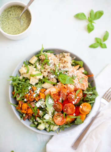 A bowl of salmon sweet potato and chimichurri salad featuring fresh greens, cherry tomatoes, sweet potatoes, corn, grains, salmon, and feta cheese sits next to a cup of green dressing, basil leaves, a napkin, and a fork on a light background.