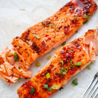 Two cooked sweet chili salmon fillets with a shiny glaze, garnished with sliced green onions, are placed on parchment paper. A fork is visible next to one fillet, and some flaky pieces of salmon are separated.
