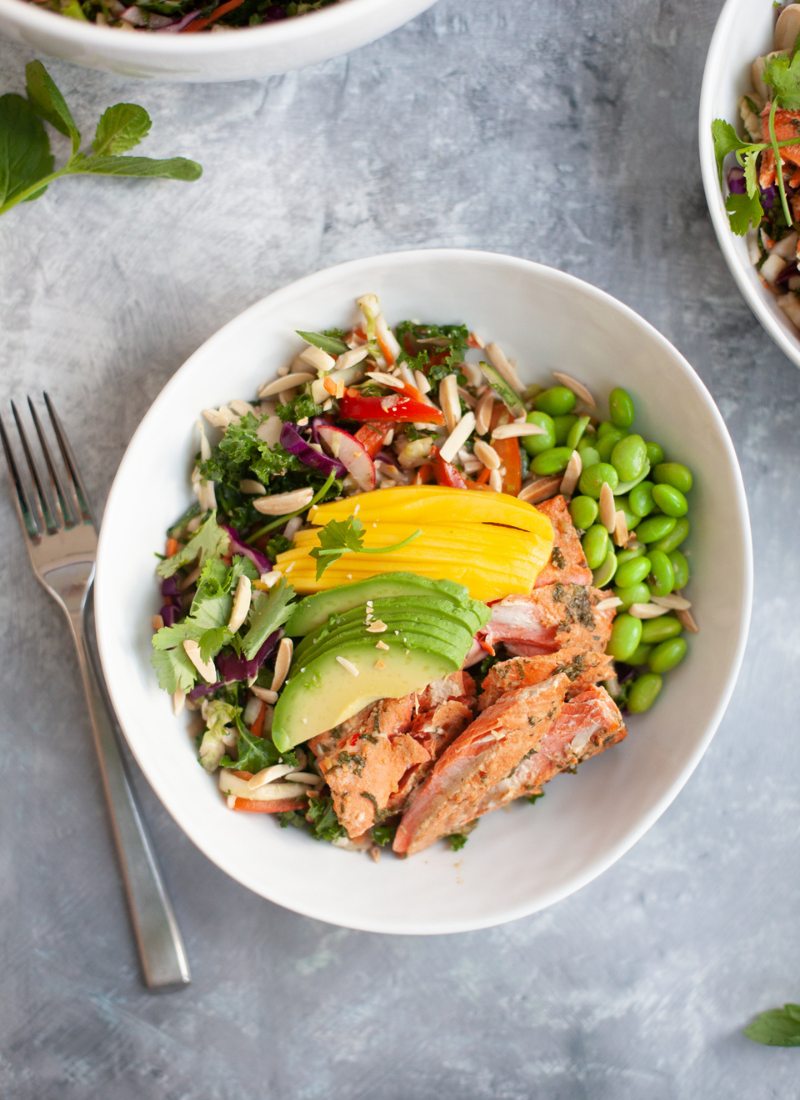 A white bowl filled with soy ginger salmon, sliced avocado, mango, edamame, greens, red onion, tomatoes, Asian slaw, and slivered almonds. A fork is placed beside the bowl on a gray surface.