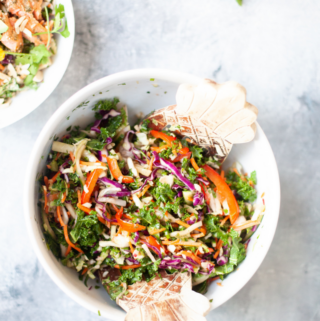 A bowl of colorful vegetable salad with shredded carrots, purple cabbage, kale, and red bell pepper, mixed with wooden salad tongs makes the perfect side for soy ginger salmon and Asian slaw. A portion of another salad bowl is visible in the corner.