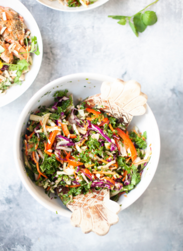 A bowl of colorful vegetable salad with shredded carrots, purple cabbage, kale, and red bell pepper, mixed with wooden salad tongs makes the perfect side for soy ginger salmon and Asian slaw. A portion of another salad bowl is visible in the corner.