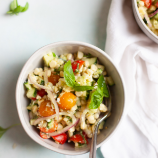 A bowl of fresh corn feta and basil salad with cherry tomatoes, red onions, basil, and diced vegetables, served with a spoon on a light surface next to a white napkin.