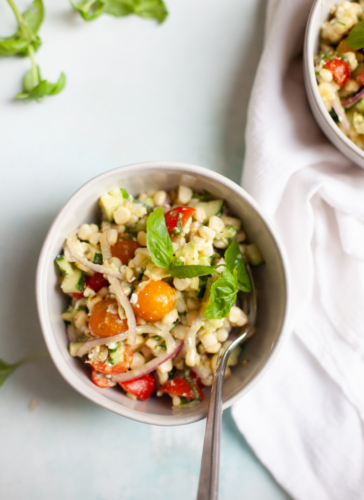 A bowl of fresh corn feta and basil salad with cherry tomatoes, red onions, basil, and diced vegetables, served with a spoon on a light surface next to a white napkin.