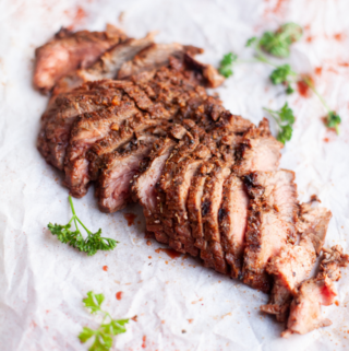 Sliced sweet garlic spiced grilled flank steak arranged on parchment paper, garnished with sprigs of parsley and scattered red spices.