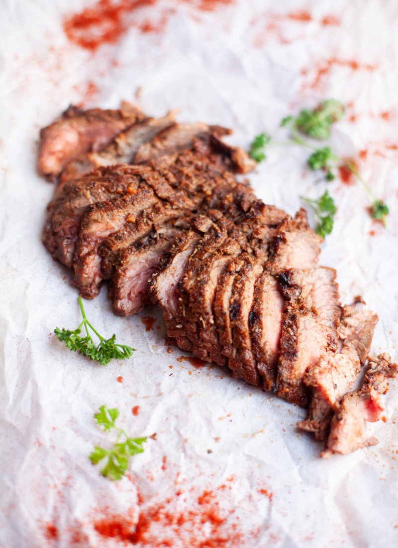 Sliced sweet garlic spiced grilled flank steak arranged on parchment paper, garnished with sprigs of parsley and scattered red spices.