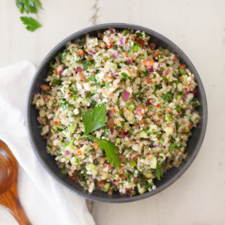 A bowl of tabouli salad with feta, brown rice, and cauliflower is beautifully garnished with parsley and sits on a white surface next to a tomato, wooden spoon, and fresh parsley sprigs.