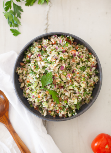 A bowl of tabouli salad with feta, brown rice, and cauliflower is beautifully garnished with parsley and sits on a white surface next to a tomato, wooden spoon, and fresh parsley sprigs.