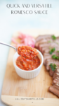 A spoonful of romesco sauce is held above a small white dish of sauce, with sliced cooked meat and herbs on a wooden board in the background. Text at the top reads, Quick and versatile romesco sauce.