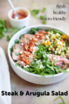 A bowl of Steak and Arugula Salad with corn, pickled onions, cheese, and red sauce. Text on image: fresh, healthy, macro-friendly. Steak & Arugula Salad. A small bowl of red sauce is in the background.
