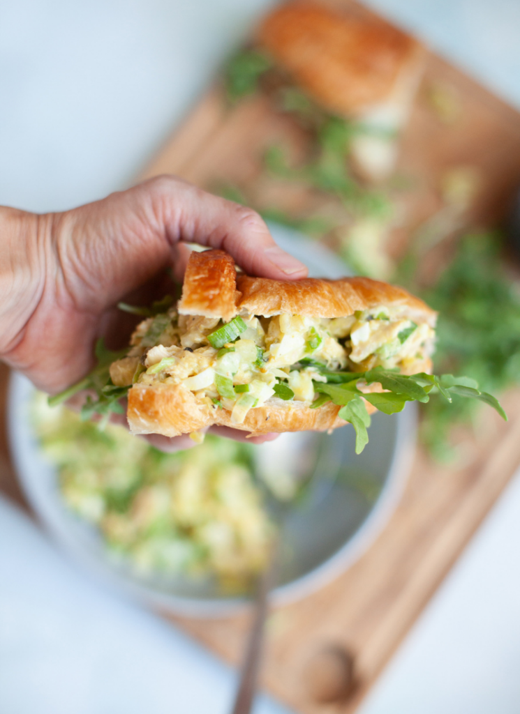 Quick and easy pickle chicken salad in croissant being help in hand. Bowl of chicken salad below sandwich on top of wooden cutting board.