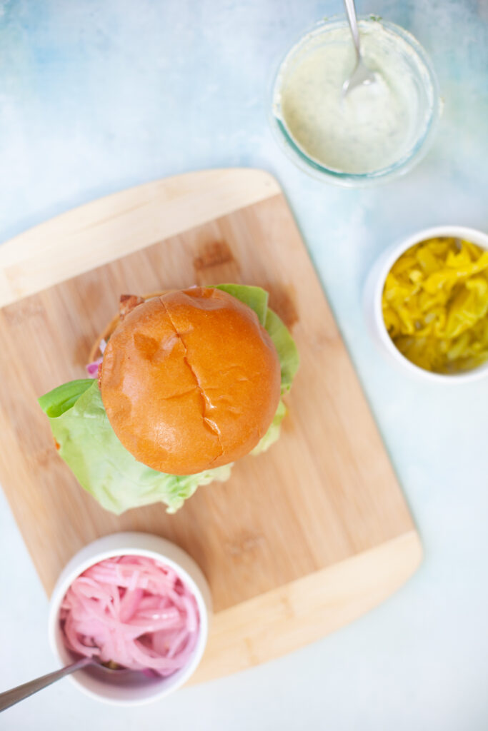 Overhead view of a Blackened Chicken Sandwich with Whipped Herb Feta on a wooden cutting board, surrounded by small bowls of pickled onions, sliced peppers, and a jar of creamy sauce on a light blue background.