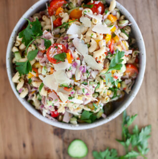A bowl of Italian Pasta Salad with ham, cherry tomatoes, red onion, cucumber, parsley, and shaved parmesan sits on a wooden table, with cucumber slices and parsley nearby.