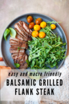 A plate with sliced Balsamic Grilled Flank Steak, fresh arugula, yellow cherry tomatoes, and basil leaves. Text reads: Easy and macro friendly Balsamic Grilled Flank Steak. A hand holds the edge of the plate.