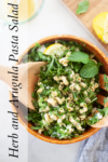 A wooden bowl filled with Herb and Arugula Pasta Salad, garnished with fresh herbs and lemon, sits alongside wooden salad tongs. Text on the left reads Herb and Arugula Pasta Salad.
