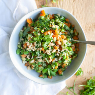 A bowl of vibrant Asian Edamame Salad, featuring chopped vegetables, herbs, and nuts, sits on a wooden table, with a white cloth and fresh greens nearby.