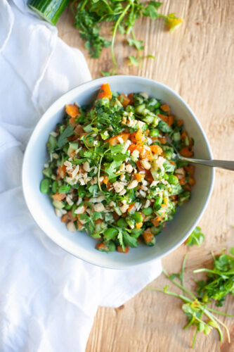 A bowl of vibrant Asian Edamame Salad, featuring chopped vegetables, herbs, and nuts, sits on a wooden table, with a white cloth and fresh greens nearby.