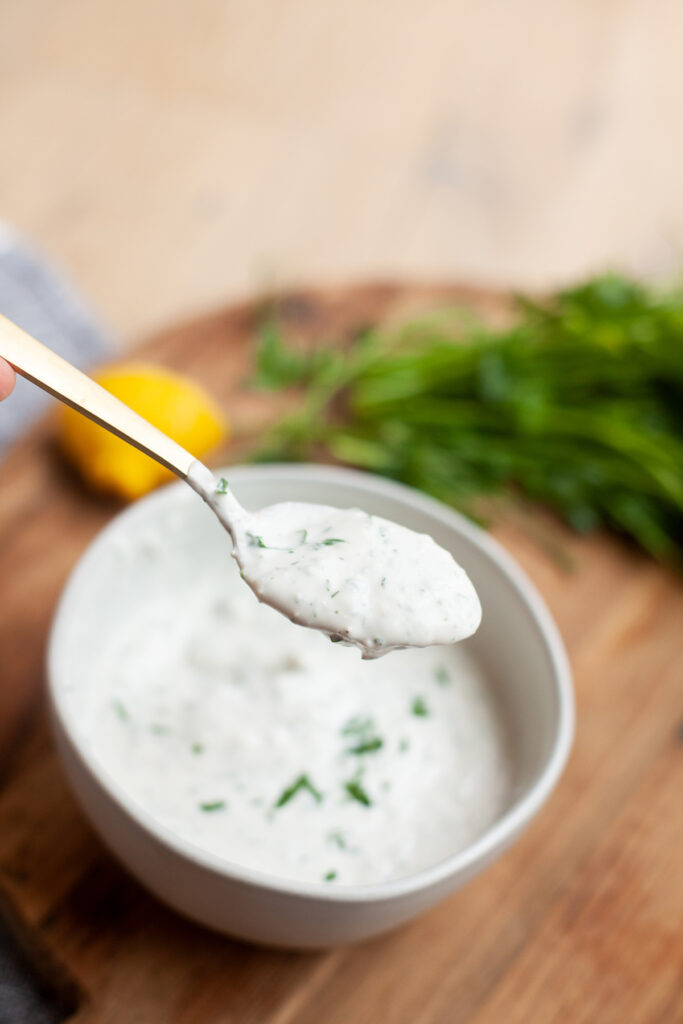A spoonful of creamy white sauce with herbs—reminiscent of the rich blue cheese dressing used for a classic Wedge Salad—is held above a bowl of the same sauce, with fresh herbs and a lemon in the blurred background.