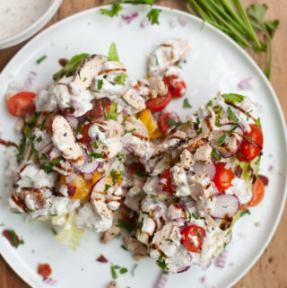 A plate with two salads, including a classic Wedge Salad with Creamy Blue Cheese Dressing, topped with grilled chicken, cherry tomatoes, onions, chopped herbs, and creamy dressing. Fresh parsley and a bowl of extra dressing are on a wooden table beside the plate.