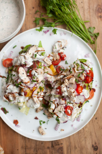 A plate with two salads, including a classic Wedge Salad with Creamy Blue Cheese Dressing, topped with grilled chicken, cherry tomatoes, onions, chopped herbs, and creamy dressing. Fresh parsley and a bowl of extra dressing are on a wooden table beside the plate.