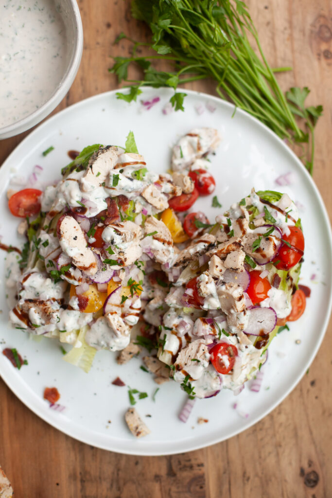 A plate with two salads, including a classic Wedge Salad with Creamy Blue Cheese Dressing, topped with grilled chicken, cherry tomatoes, onions, chopped herbs, and creamy dressing. Fresh parsley and a bowl of extra dressing are on a wooden table beside the plate.