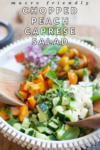 A bowl of vibrant Chopped Peach Caprese Salad features crisp cucumbers, juicy tomatoes, sweet peaches, onions, mozzarella, fresh basil, and a drizzle of pesto. Two wooden spoons rest nearby for easy serving. Text over image reads Macro Friendly Chopped Peach Caprese Salad.
