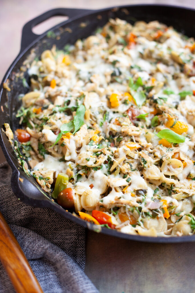 A black skillet filled with a cheesy One Pan Garden Veggie Orecchiette bake, featuring shell pasta, chopped vegetables, herbs, and melted cheese, sits on a gray cloth next to a wooden-handled utensil.