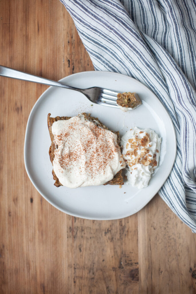 A slice of cake topped with cream and cinnamon sits on a white plate, alongside whipped cream and chopped nuts—reminiscent of the cozy flavors found in pumpkin pie baked protein oat bars. A fork rests on the plate, and a striped cloth is nearby on a wooden surface.