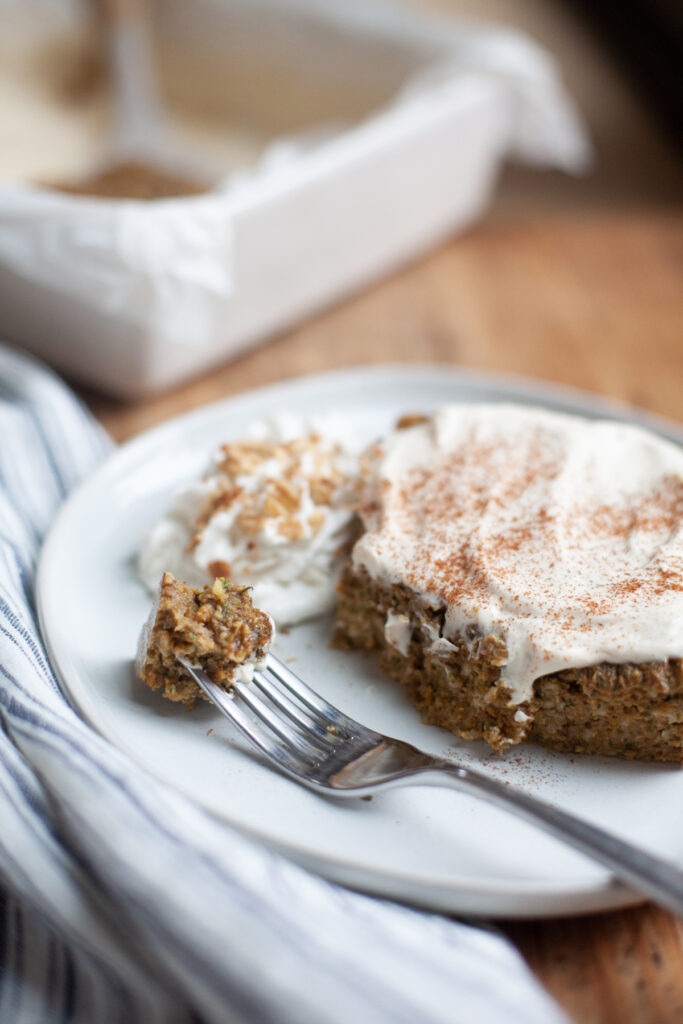 A slice of frosted cake with a bite taken out sits on a white plate next to a dollop of whipped cream; a fork holds a bite of the cake, reminiscent of indulging in pumpkin pie baked protein oat bars. A baking dish is blurred in the background.