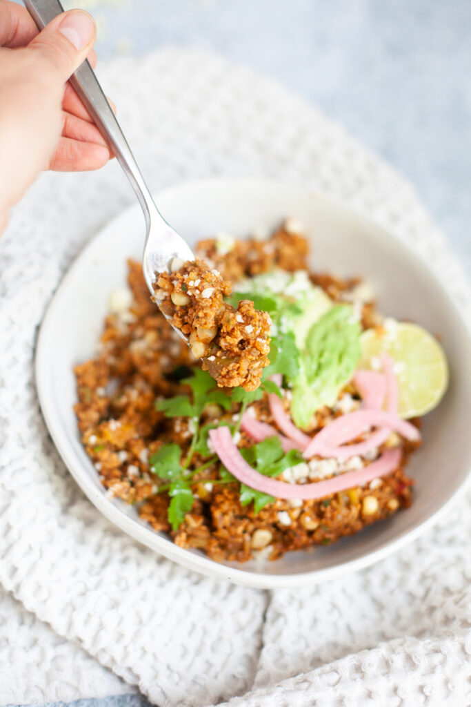 A hand holds a fork with seasoned quinoa and Mexican Cauliflower Fried Rice over a bowl topped with avocado, pickled onions, fresh cilantro, cheese crumbles, and a lime wedge on a textured white napkin.