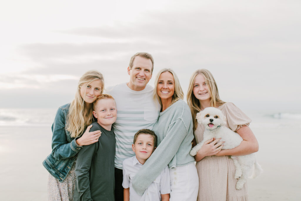A family of six, including a small white dog, poses closely together on a beach. The adults and children are smiling, dressed in light-colored casual clothes, with the ocean and sky in the background—capturing a moment that feels right out of an About Lillie Eats and Tells story, where wholesome family moments and beautiful scenery come together seamlessly.