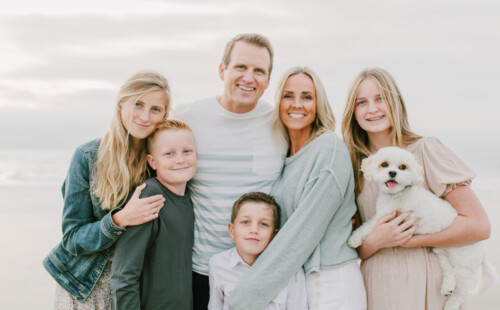 A family of six, including a small white dog, poses closely together on a beach. The adults and children are smiling, dressed in light-colored casual clothes, with the ocean and sky in the background—capturing a moment that feels right out of an About Lillie Eats and Tells story, where wholesome family moments and beautiful scenery come together seamlessly.