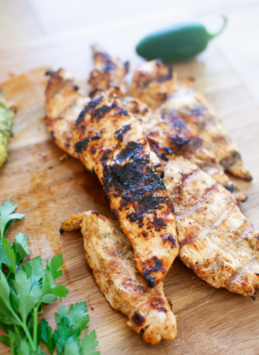 Honey chipotle grilled chicken breast strips with char marks on a wooden cutting board, garnished with fresh parsley and a jalapeño pepper in the background.