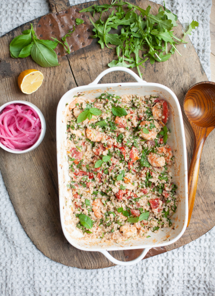 A white baking dish with baked feta cauliflower rice with shrimp and couscous salad sits on a wooden board, surrounded by fresh herbs, half a lemon, a bowl of pickled onions, and a wooden spoon.