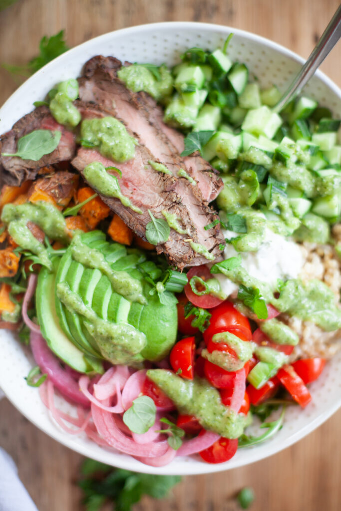 A colorful bowl with sliced steak, avocado, cucumbers, grape tomatoes, red onions, sweet potatoes, greens, and a vibrant dairy-free green sauce drizzled on top.