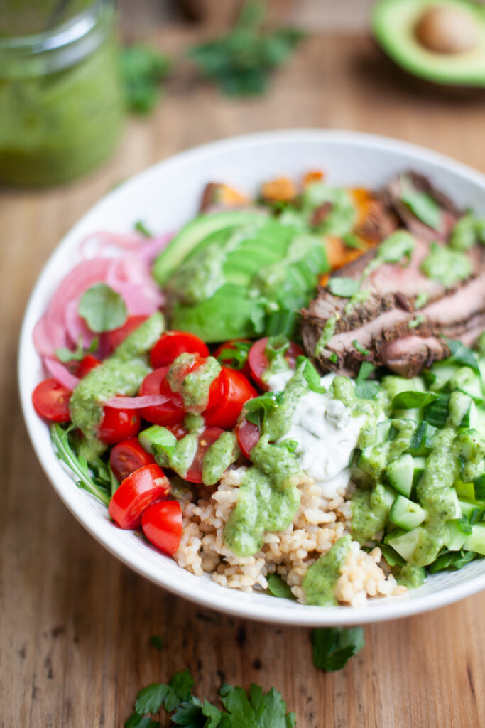 A bowl with sliced steak, brown rice, cherry tomatoes, cucumber, avocado, greens, pickled onions, and a vibrant dairy-free green sauce on top, served on a wooden table.