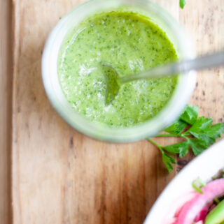 A jar of dairy-free green sauce with a spoon in it sits on a wooden surface, near fresh parsley and a partial bowl of salad with red onions.