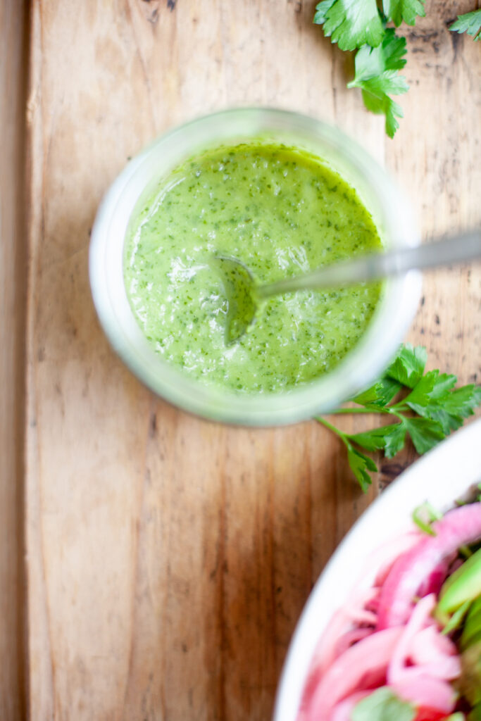A jar of dairy-free green sauce with a spoon in it sits on a wooden surface, near fresh parsley and a partial bowl of salad with red onions.