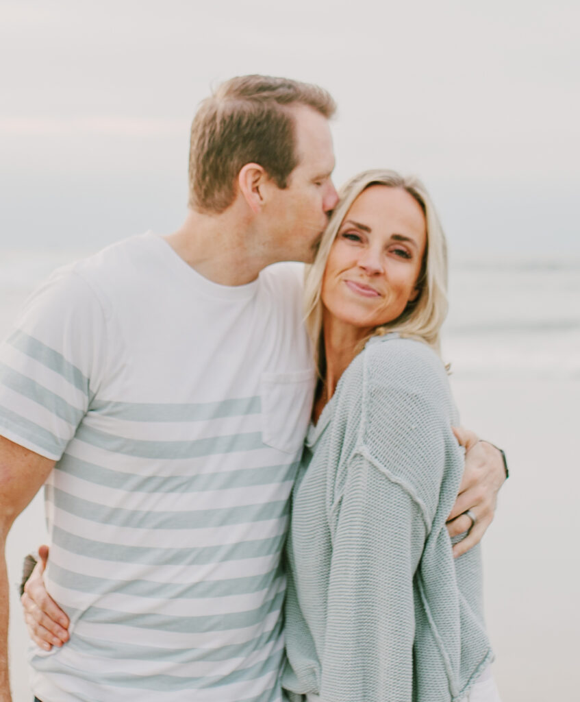 Man kisses woman on the forehead while hugging her on a beach. Both are smiling, dressed casually, with the ocean and cloudy sky in the background—reminiscent of sharing happy moments after enjoying macro friendly dinner recipes together.