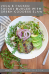 White bowl with a Veggie Packed Turkey Burger, sliced avocado, red onion, arugula, vibrant Green Goddess Slaw, and dressing on a wood table. A striped cloth and fresh greens surround the bowl. Text overlay describes the meal.
