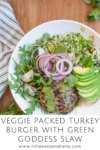 A bowl with a Veggie Packed Turkey Burger with Green Goddess Slaw, sliced avocado, arugula, slaw, red onion, and cucumber, set on a wooden table next to a striped napkin. Text below reads: Veggie Packed Turkey Burger with Green Goddess Slaw.
